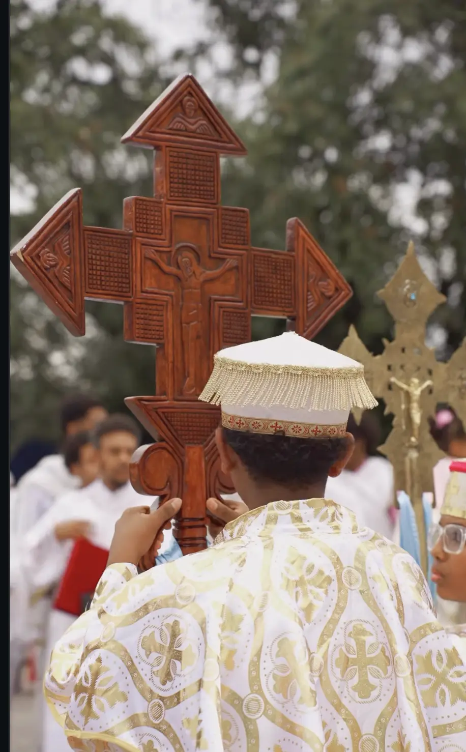 Deacon in gold vestments carrying a carved wooden processional cross during an outdoor church celebration